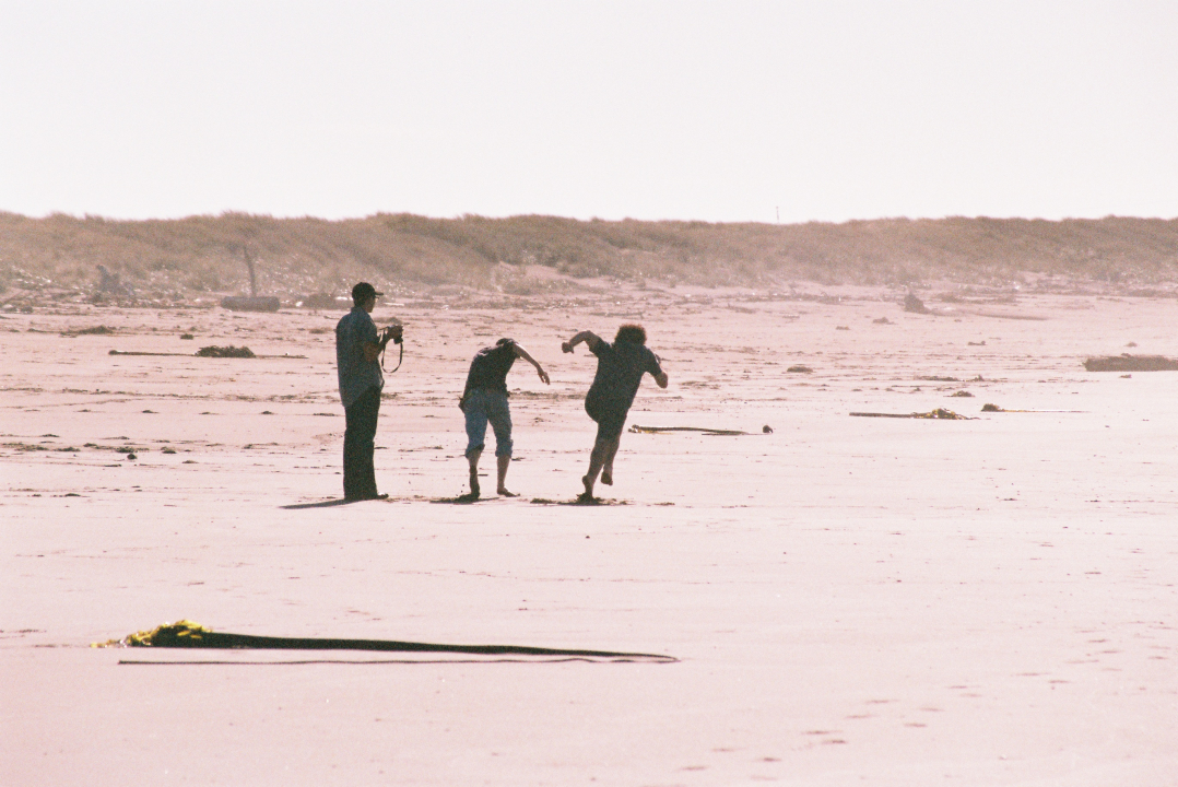 three wolves (co-opers who live at Wolf) at the beach, two running barefoot with their pants legs rolled up, one standing with a camera