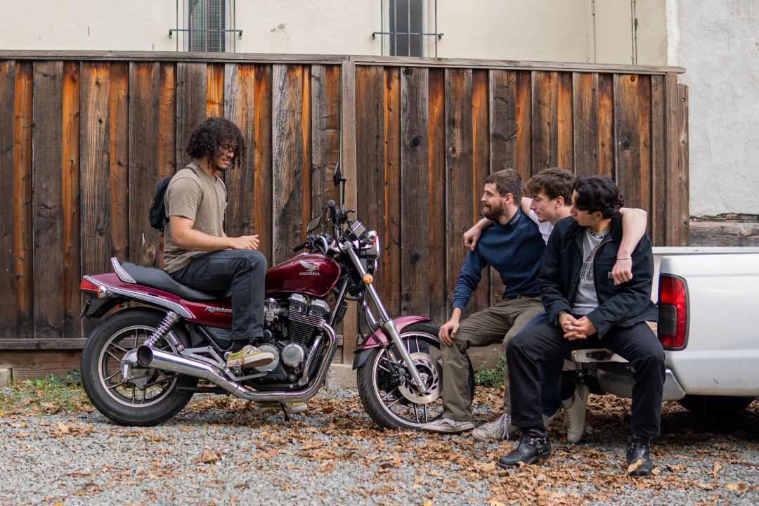a wolf (a co-oper who lives at Wolf) sitting on a motorcycle talking to three more who are sitting on the tailgate of a pickup truck