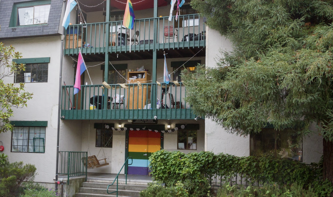 Oscar Wilde Entrance. A multi-level building features balconies draped with Pride flags and a rainbow-painted front door. Surrounded by trees and shrubs, the entrance looks welcoming and inclusive.