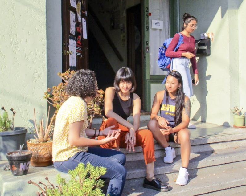 Girls sitting outside of Hoyt Hall. Four people sit on the steps of a building, talking and laughing in the sunshine, surrounded by potted plants.
