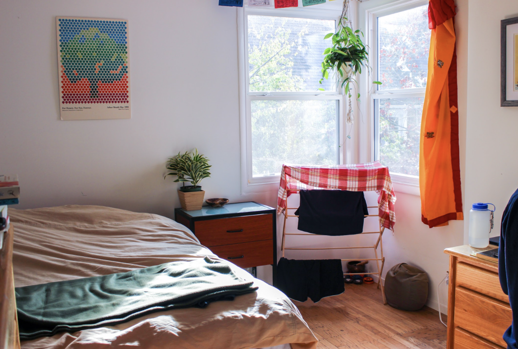 Room in a Co-op. A bright, sunlit bedroom with white walls, a bed dressed in neutral linens, and a wooden nightstand holding a small potted plant. A clothes drying rack draped with red-checkered fabric sits by the window, which also features a hanging plant and an orange cloth on the wall.