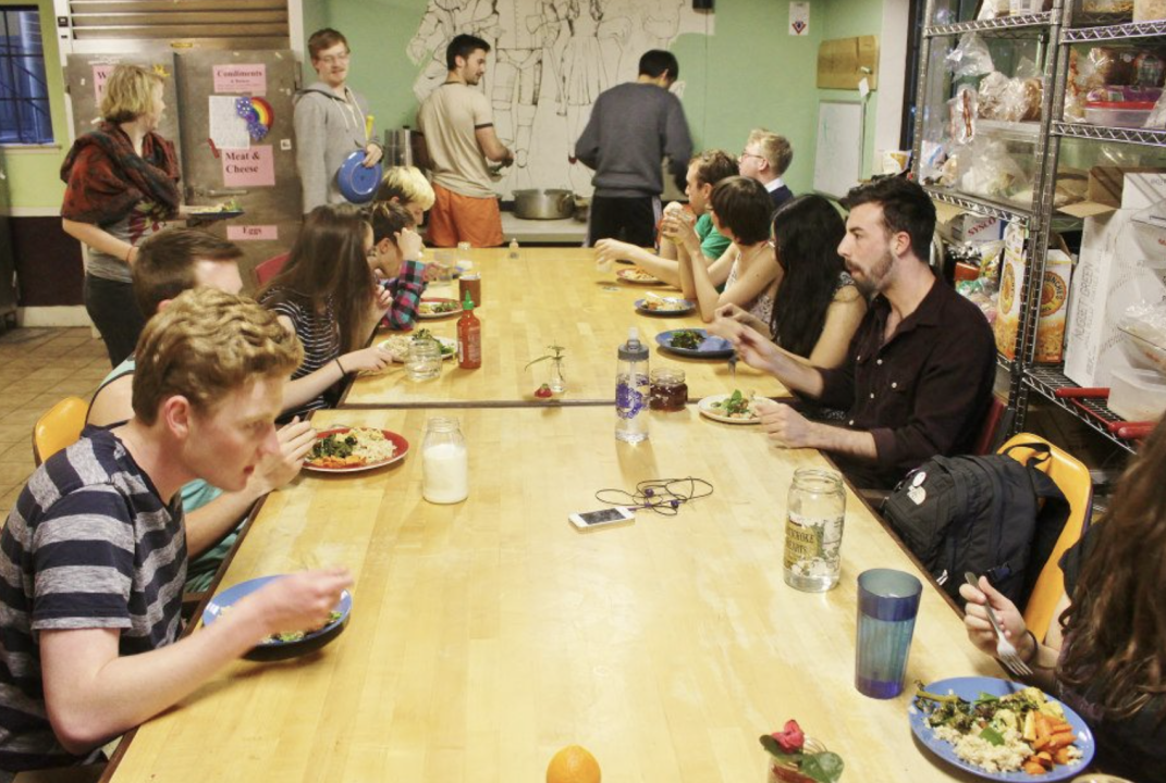 People eating food in a co-op. A long wooden table filled with people enjoying a meal together, chatting and sharing food in a relaxed group setting.
