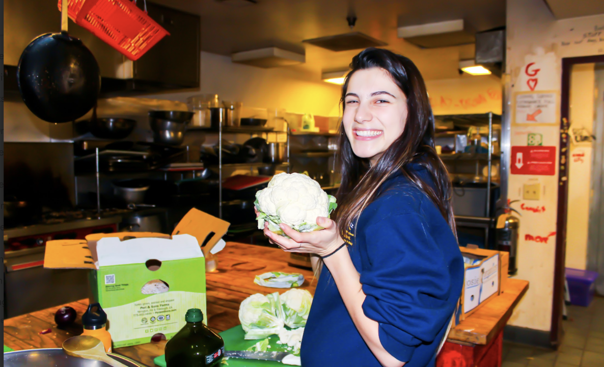 Person holding cabbage in a kitchen. A smiling individual stands in a co-op kitchen, proudly holding up a large white head of cabbage (or cauliflower).