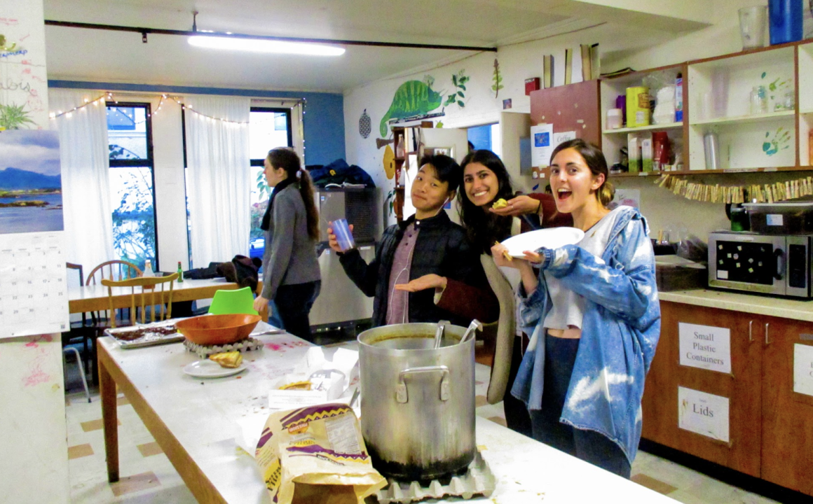 People cooking in a co-op. A group gathers in a communal kitchen, preparing and serving food around a long counter. Large pots and fresh ingredients highlight the shared, collaborative atmosphere.