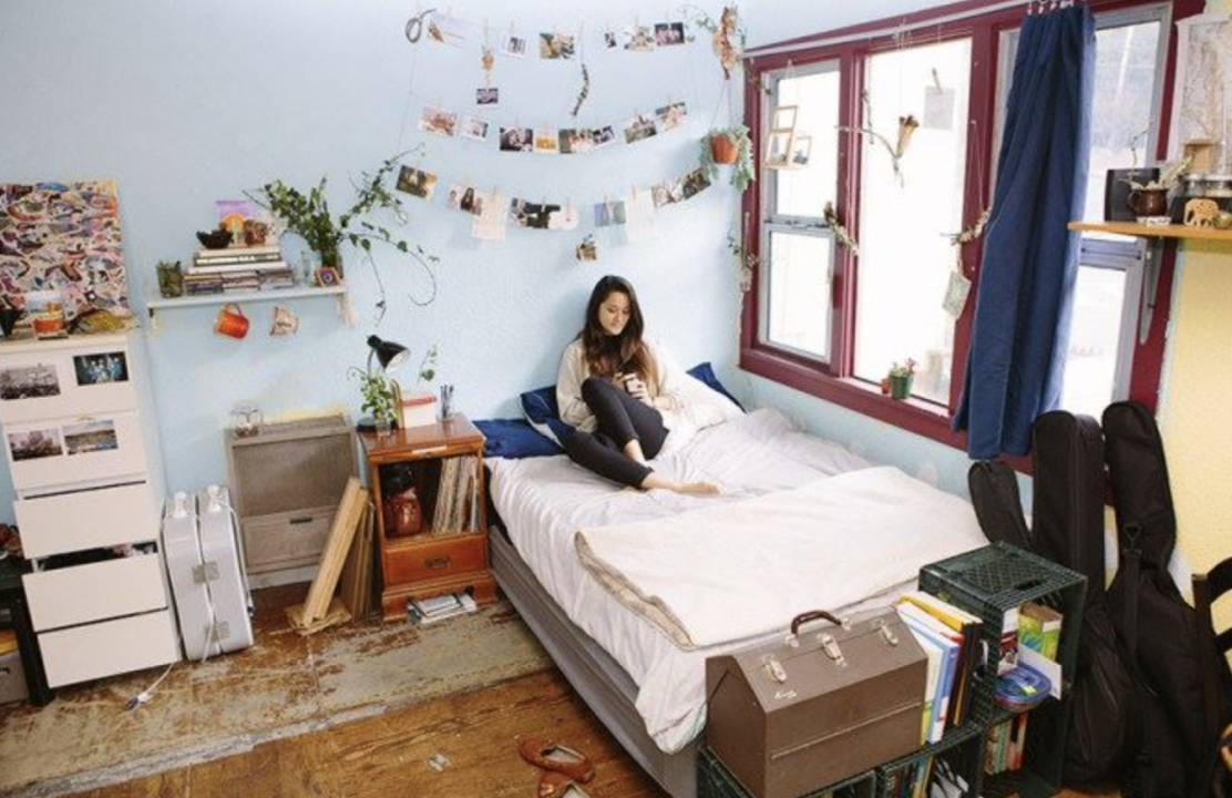 Woman sitting on bed in room in a co-op. A bright bedroom featuring a bed by the window and walls adorned with photos. A person sits on the bed with a laptop, adding a personal, lived-in feel.