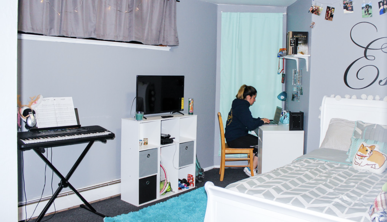 Person studying in room at co-op. A neat bedroom with gray walls, a white bed, and a small desk in the corner. A person studies at the desk while a keyboard stands nearby, reflecting a creative, personalized space.