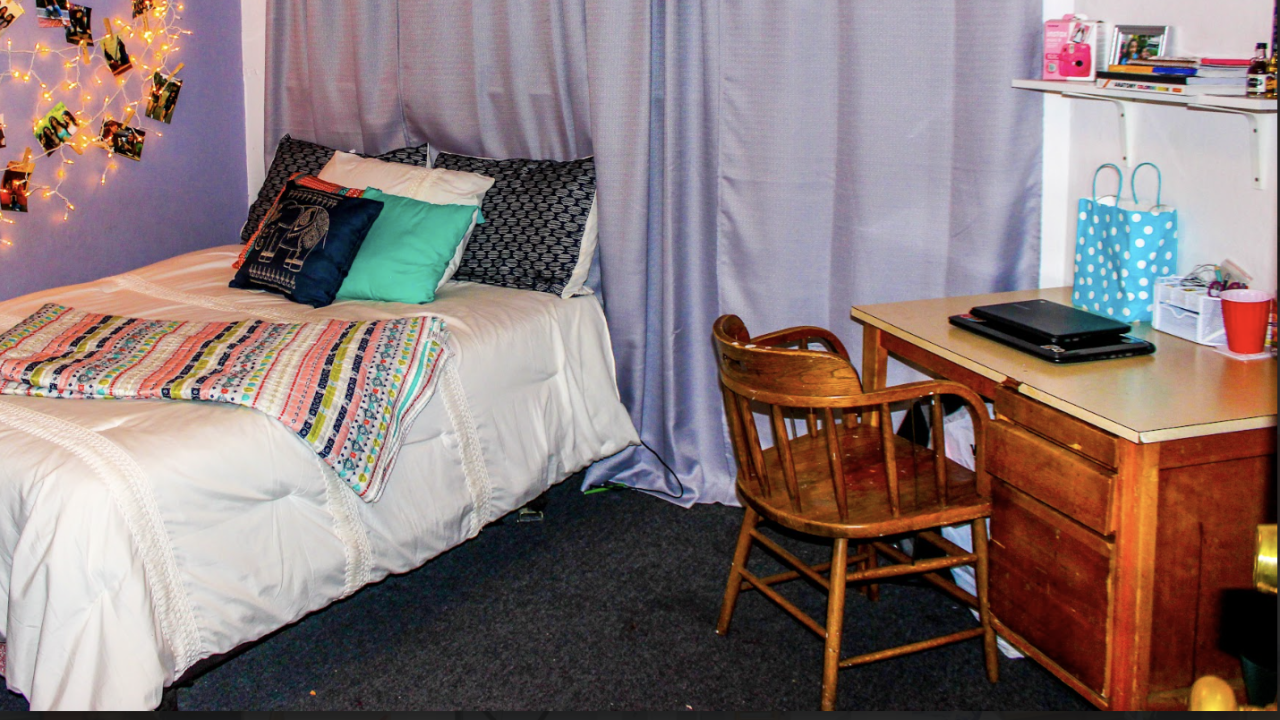Room in a co-op with bed and table. A cozy bedroom with a bed, decorative pillows, and a wooden desk next to a purple curtain. The soft color scheme and simple decor create a calm, inviting space.