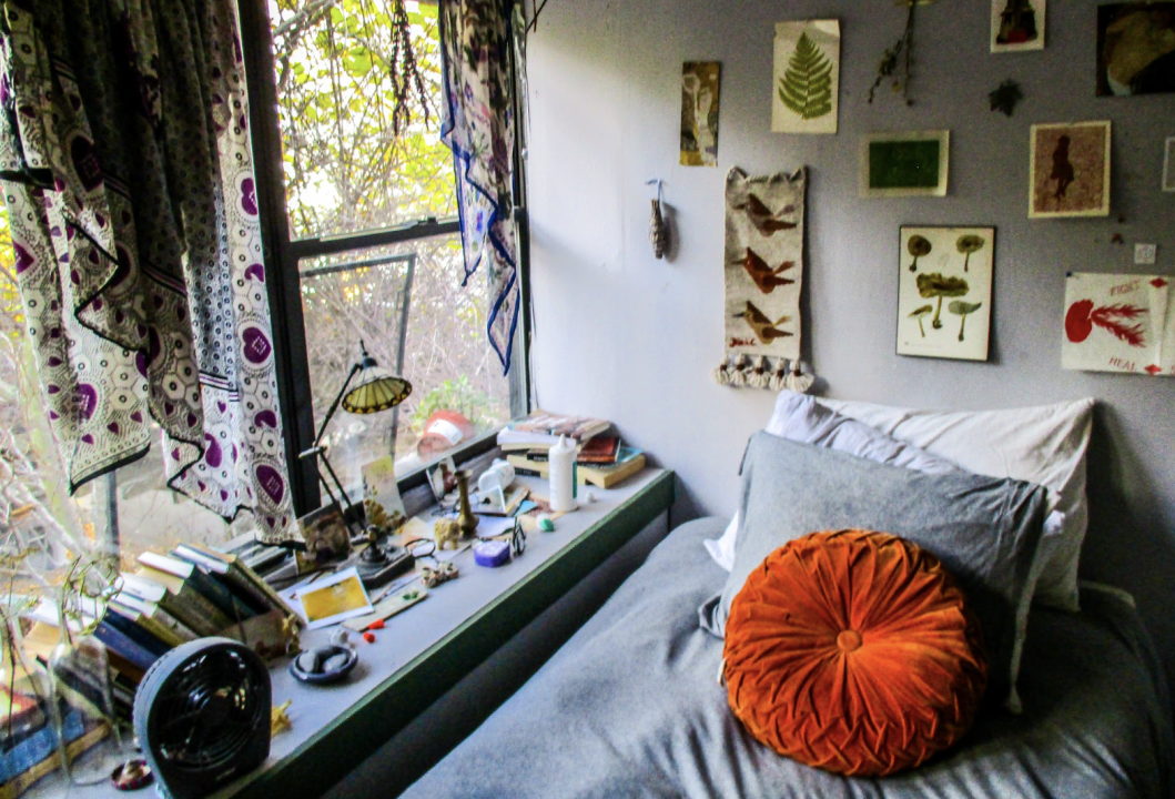 Room in a house. A bed sits by a window adorned with nature prints and pressed leaves on the wall. The windowsill is lined with small items, and an orange accent pillow adds a warm touch.