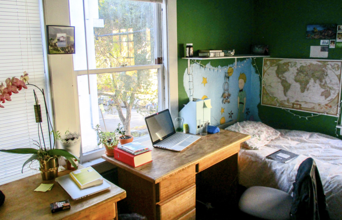 Room in a house. A bedroom with a green accent wall, a wooden desk by a window, and a bed in the corner. Maps and artwork decorate the walls, and a laptop is open on the desk.
