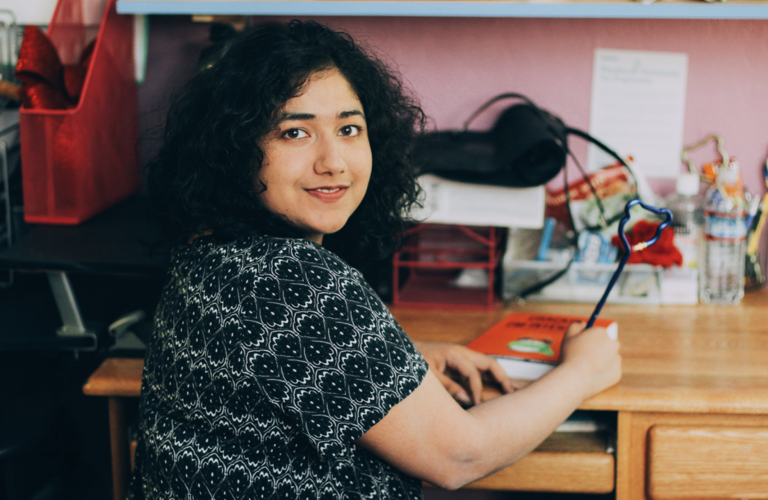 A woman with shoulder-length, dark, curly hair is seated at a wooden desk. She’s turned slightly toward the camera, looking over her shoulder with a gentle smile. She’s wearing a short-sleeved patterned top and appears to be writing in a notebook or journal. The desk is scattered with small items and stationery supplies, and the background includes a pink wall and shelving.